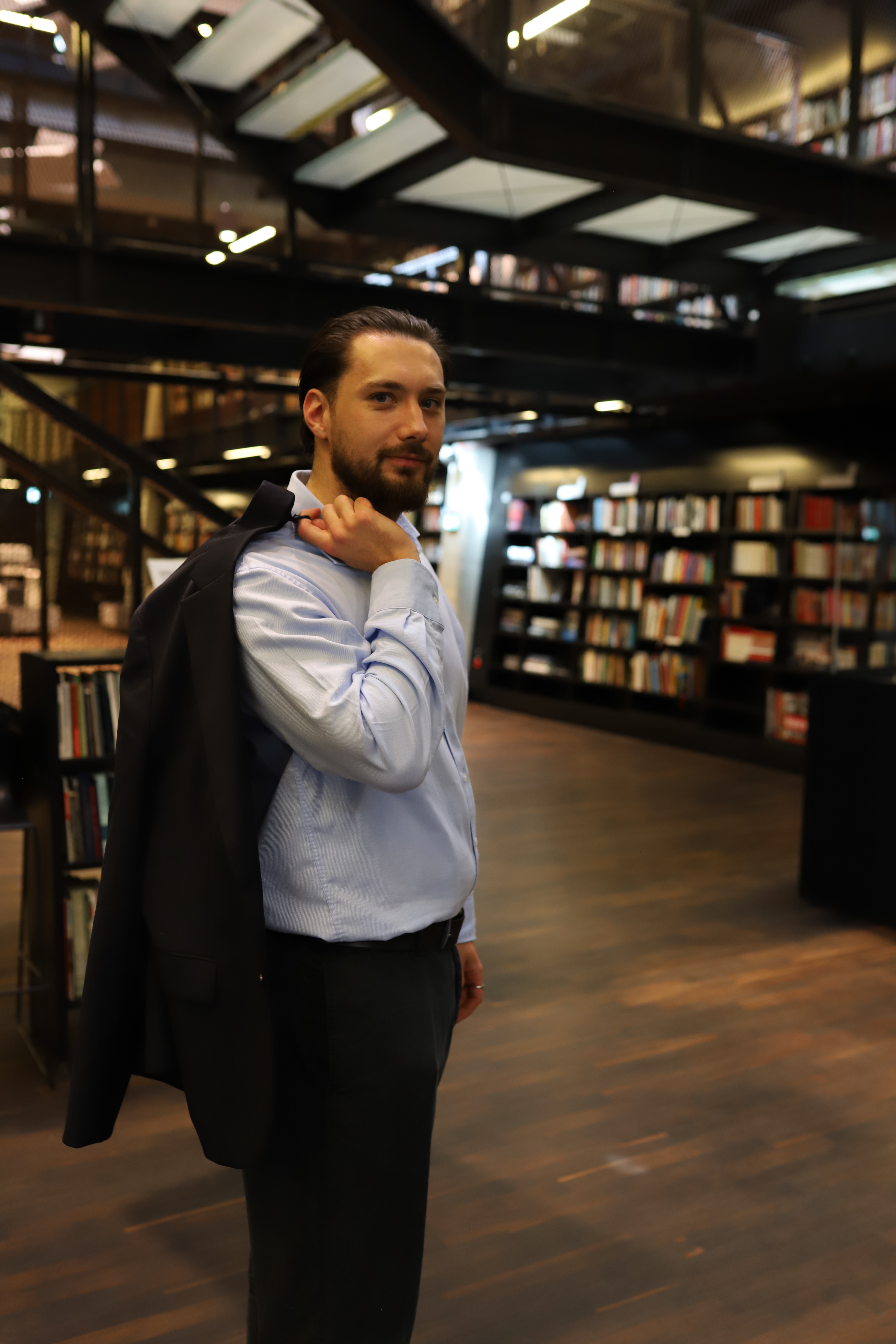 A man with dark hair and a beard, styled back, stands in a well-lit library with stacks of books and wood floors. He wears...